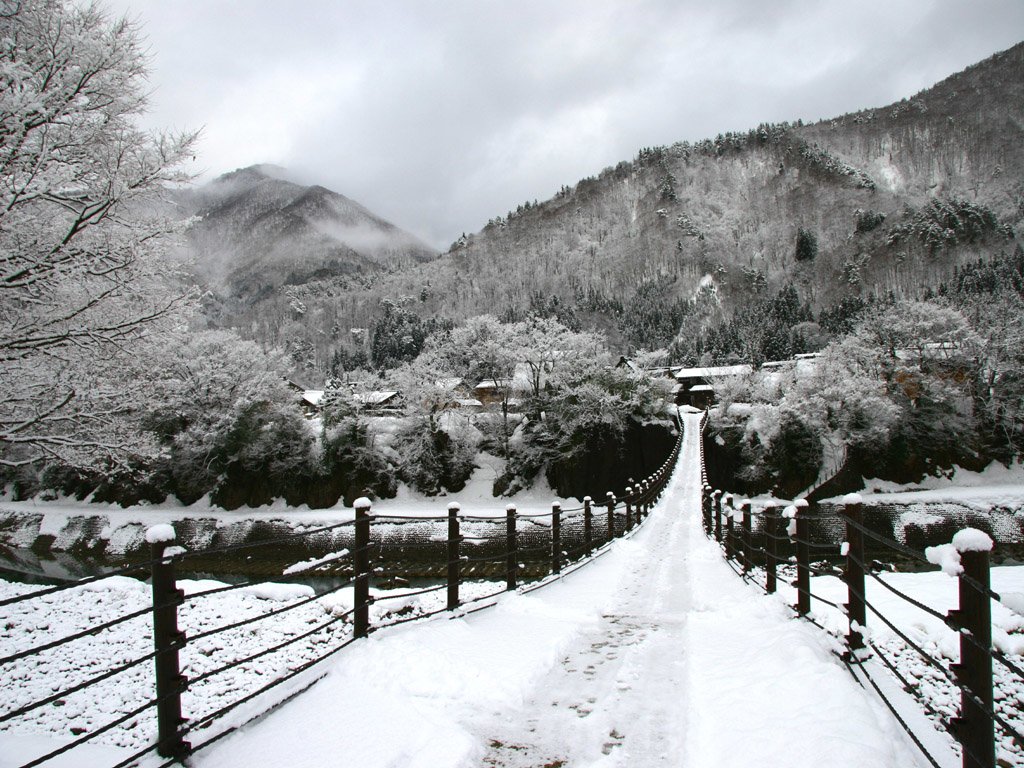 A serene winter landscape featuring a snow-covered path leading across a bridge, surrounded by frosted trees and mountains under a cloudy sky.