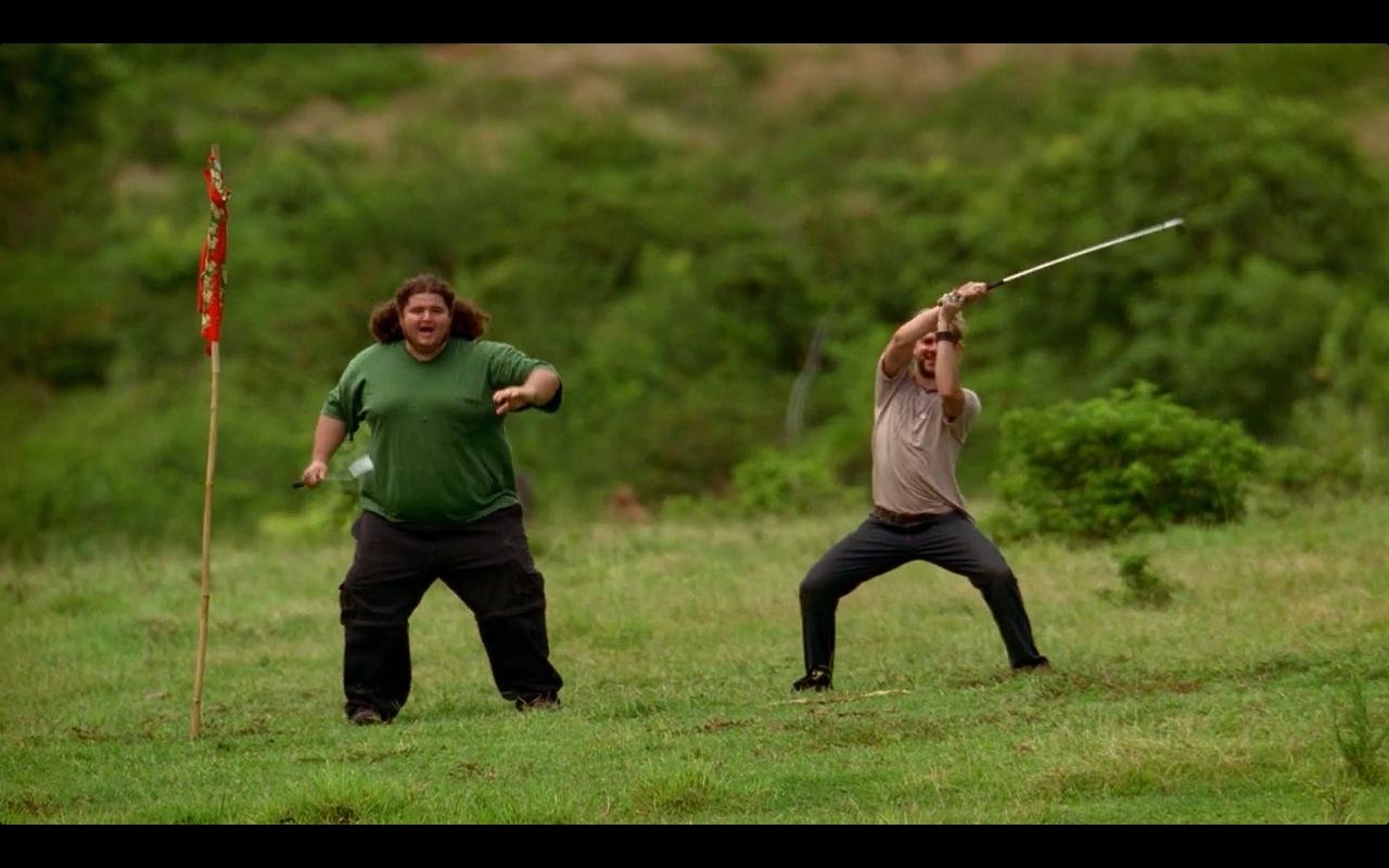 Scene from the TV show Lost: two men on a grassy field, one in green cheering beside a flag, the other poised to throw a spear-like stick.