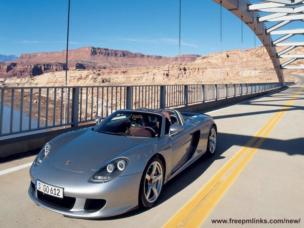A silver Porsche Carrera GT parked on a scenic bridge, with a dramatic desert landscape in the background, showcasing its sleek design and performance.