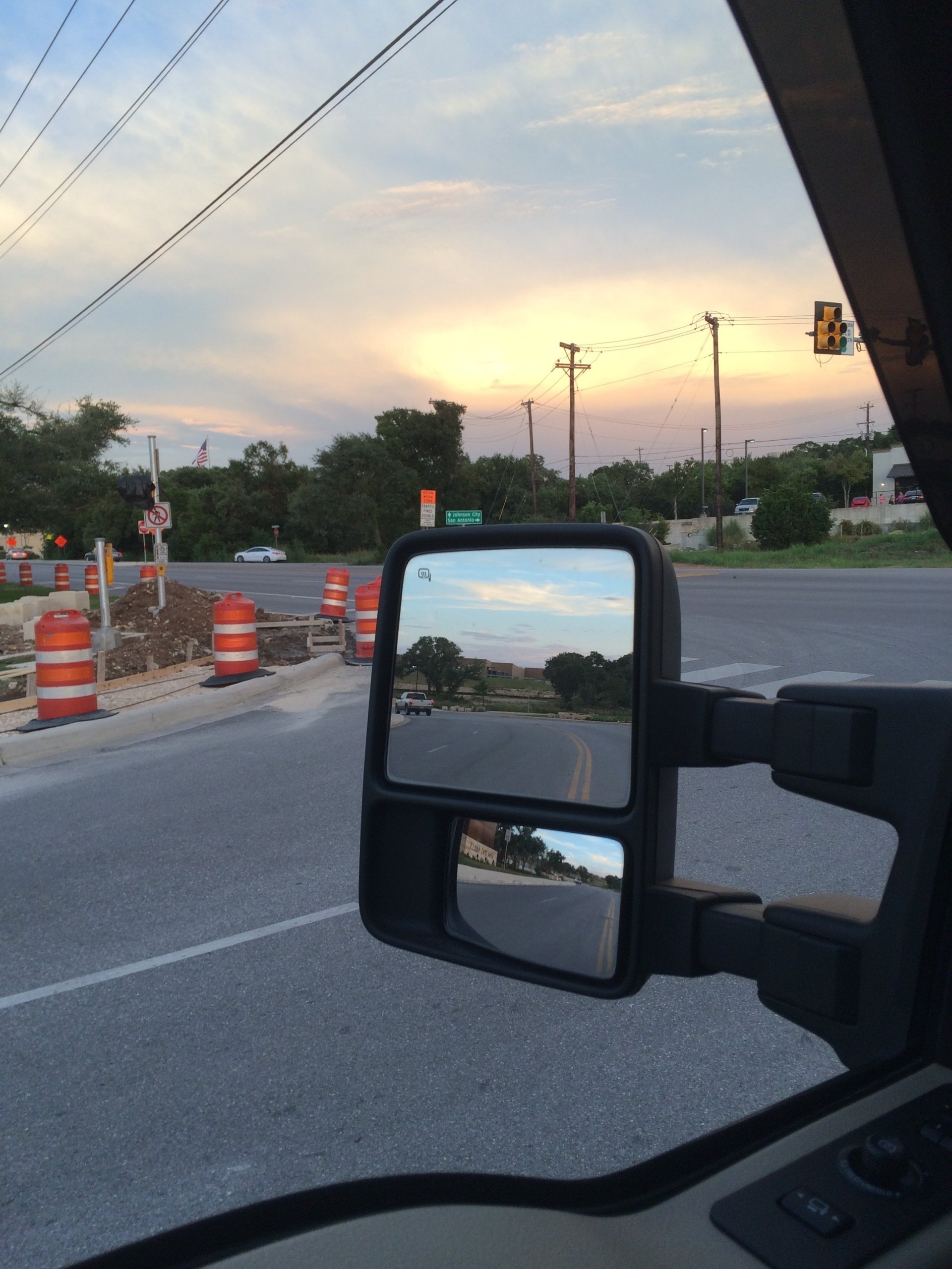 A view from a vehicle's side mirror shows a road under a colorful sunset, with construction barriers and traffic lights indicating ongoing road work.