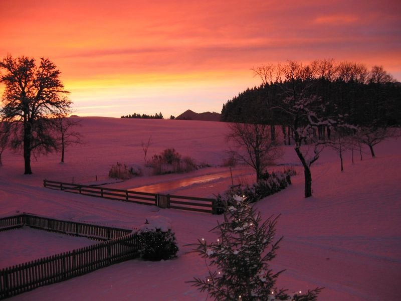 A stunning sunset casts vibrant colors over a snowy landscape, with a tranquil pond reflecting the sky and trees silhouetted against the colorful horizon.