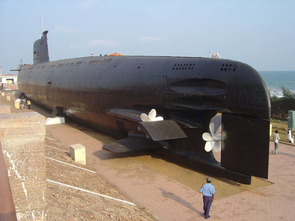 A black military submarine on display, showcasing its large propellers and sleek design, set against a backdrop of the sky and ocean. A visitor walks nearby.