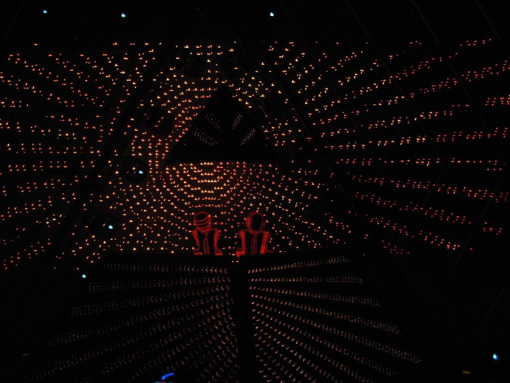 A dimly lit stage features two figures in iconic helmets, surrounded by mesmerizing light patterns, creating a vibrant atmosphere for a Daft Punk music performance.
