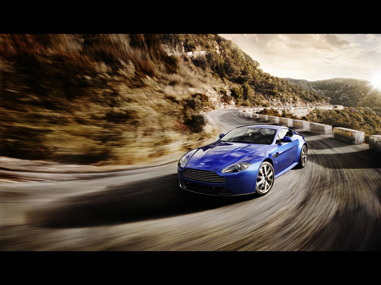 Blue Aston Martin Vantage convertible racing along a winding mountain road at sunset, motion blur emphasizing its sleek lines and sporty stance.