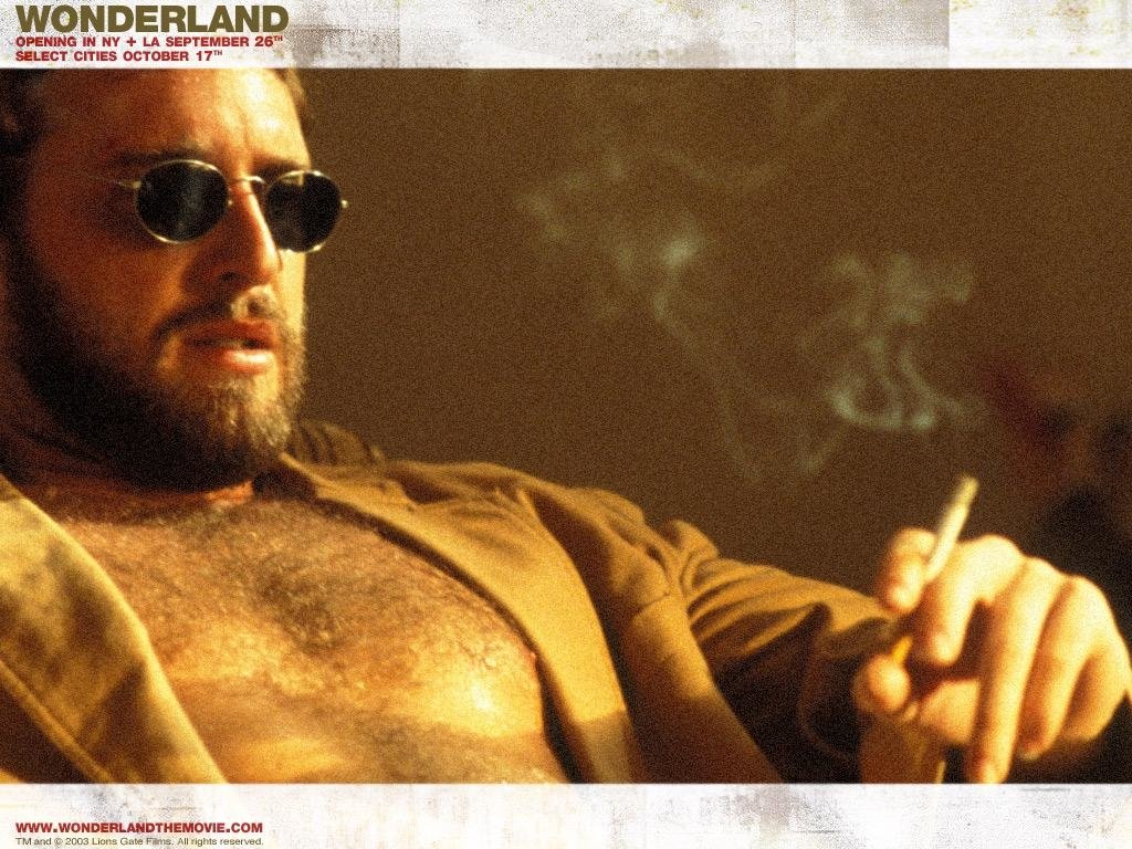 Bearded man in sunglasses, chest exposed, smoking a cigarette in a dim room — scene from the movie Wonderland (2003).