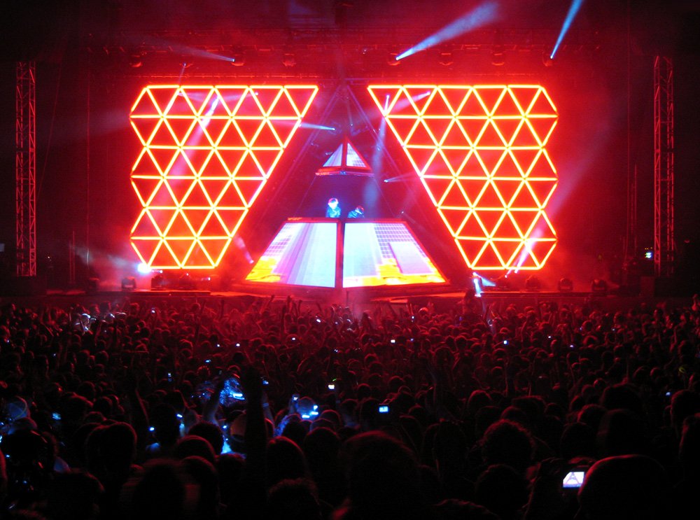 A vibrant concert scene featuring a large crowd in front of an impressive Daft Punk stage setup, illuminated by striking red triangular lights.