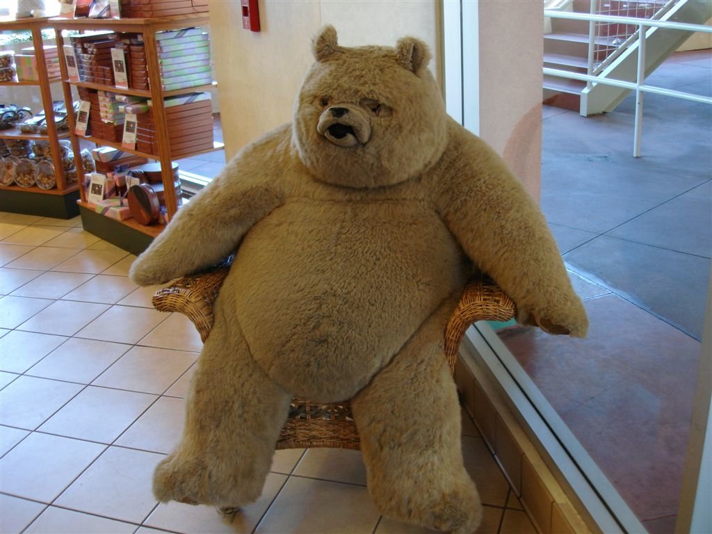 A large, man-made teddy bear sits comfortably on a chair, showcasing its plush, stuffed animal design in a bright, welcoming space.