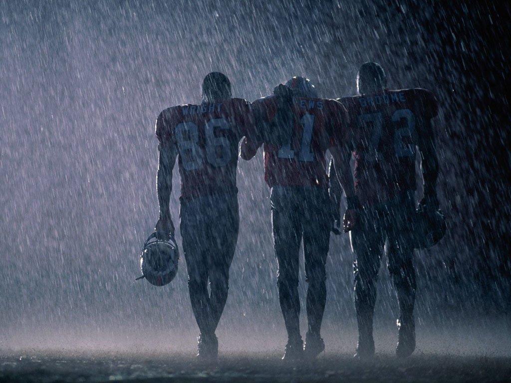 Three football players walk shoulder to shoulder in heavy rain, showcasing camaraderie and resilience in the face of challenging weather during a sports event.