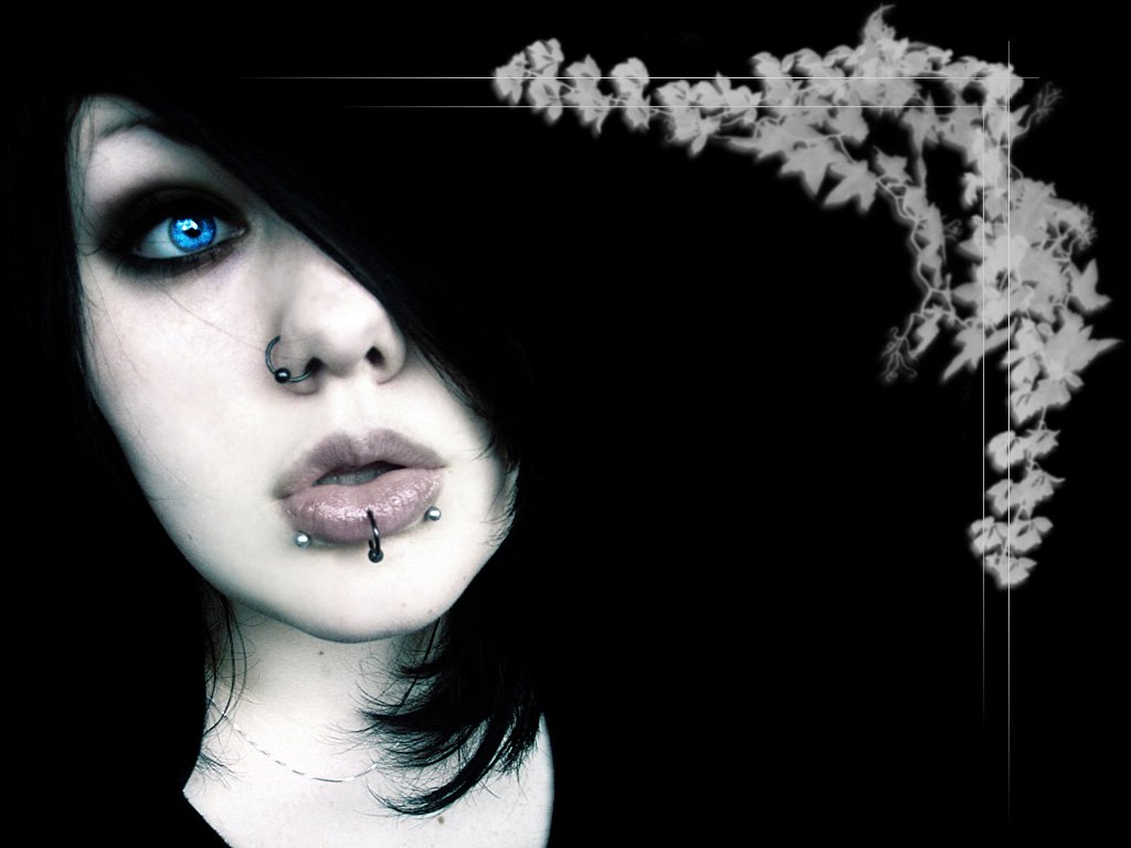 A woman with striking blue eyes and dark hair partially obscuring her face showcases multiple piercings, giving her a spooky, edgy appearance against a black background.