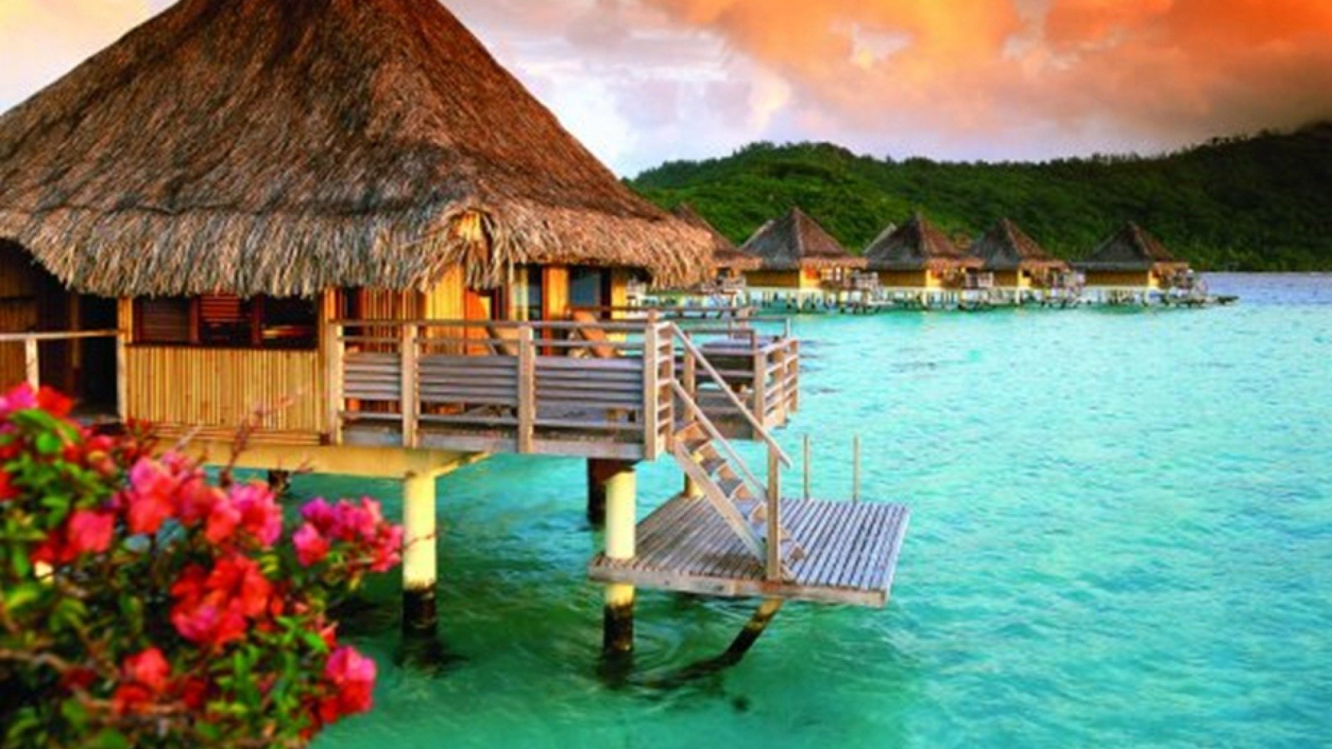 A serene holiday scene featuring overwater huts with thatched roofs, vibrant flowers, and turquoise water under a colorful sunset sky.