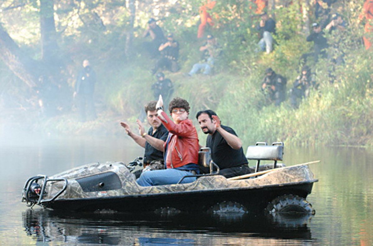 Three characters from the TV show Trailer Park Boys wave from a camouflaged boat in a misty lake surrounded by trees, capturing a humorous moment in their misadventures.