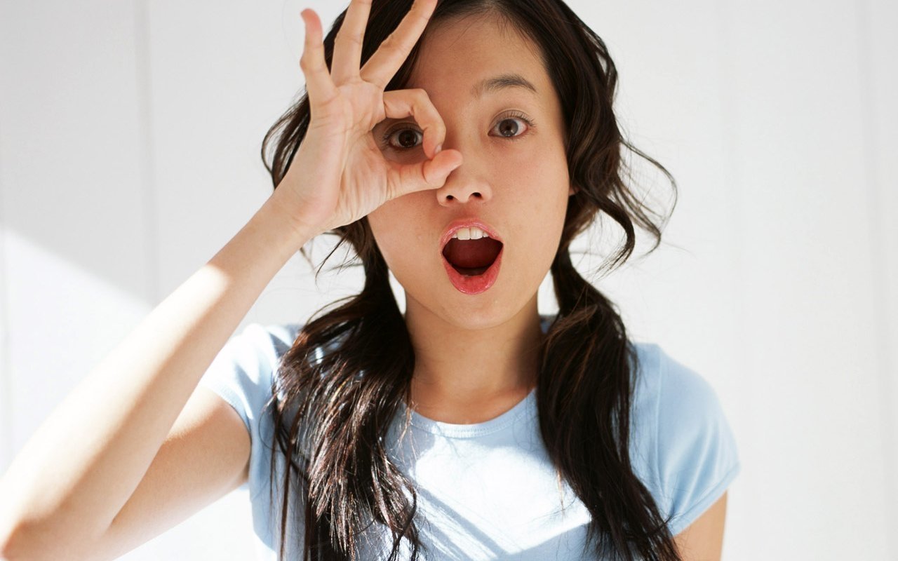 A woman playfully poses with her hand making an 'O' shape, mouth open in a surprised expression. Her long dark hair cascades in soft waves, exuding a sensual and joyful vibe.