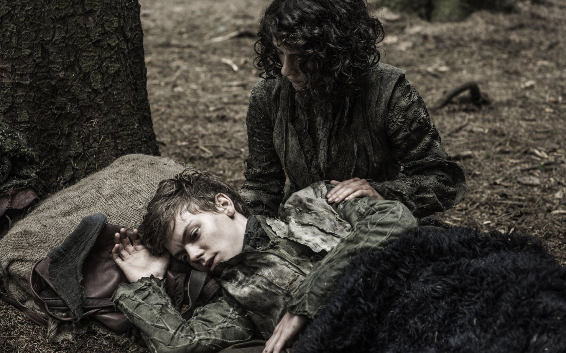Meera Reed watches over the sleeping Jojen Reed, curled on a cloak in a bleak forest scene from Game of Thrones.