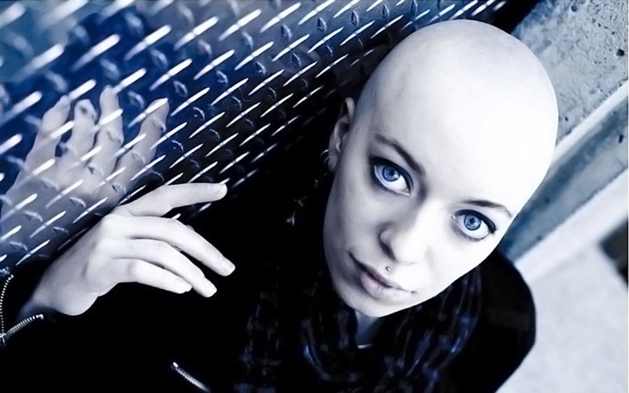 A woman with piercing blue eyes and a shaved head gazes directly at the camera, her reflection visible on a metal surface, creating a striking visual contrast.