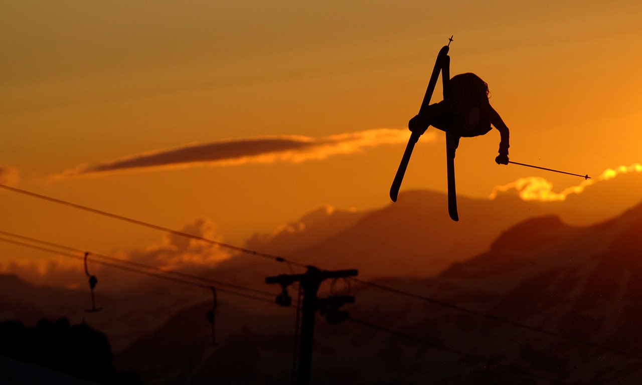 A skier performs an impressive jump against a vibrant sunset, capturing the thrill and energy of the sport amidst a backdrop of majestic mountains.