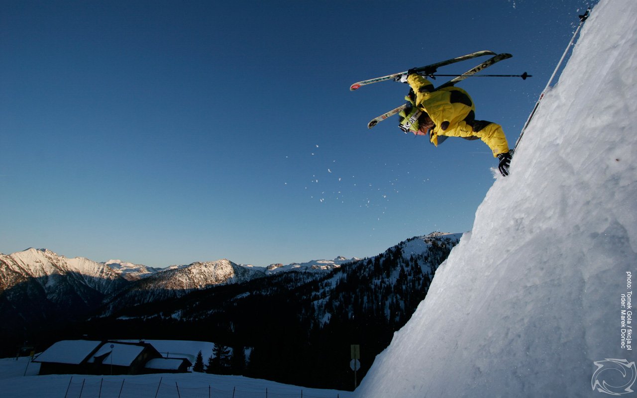 A skier in bright yellow gear gracefully launches off a snow-covered slope, showcasing athleticism against a stunning mountain backdrop during a clear day.