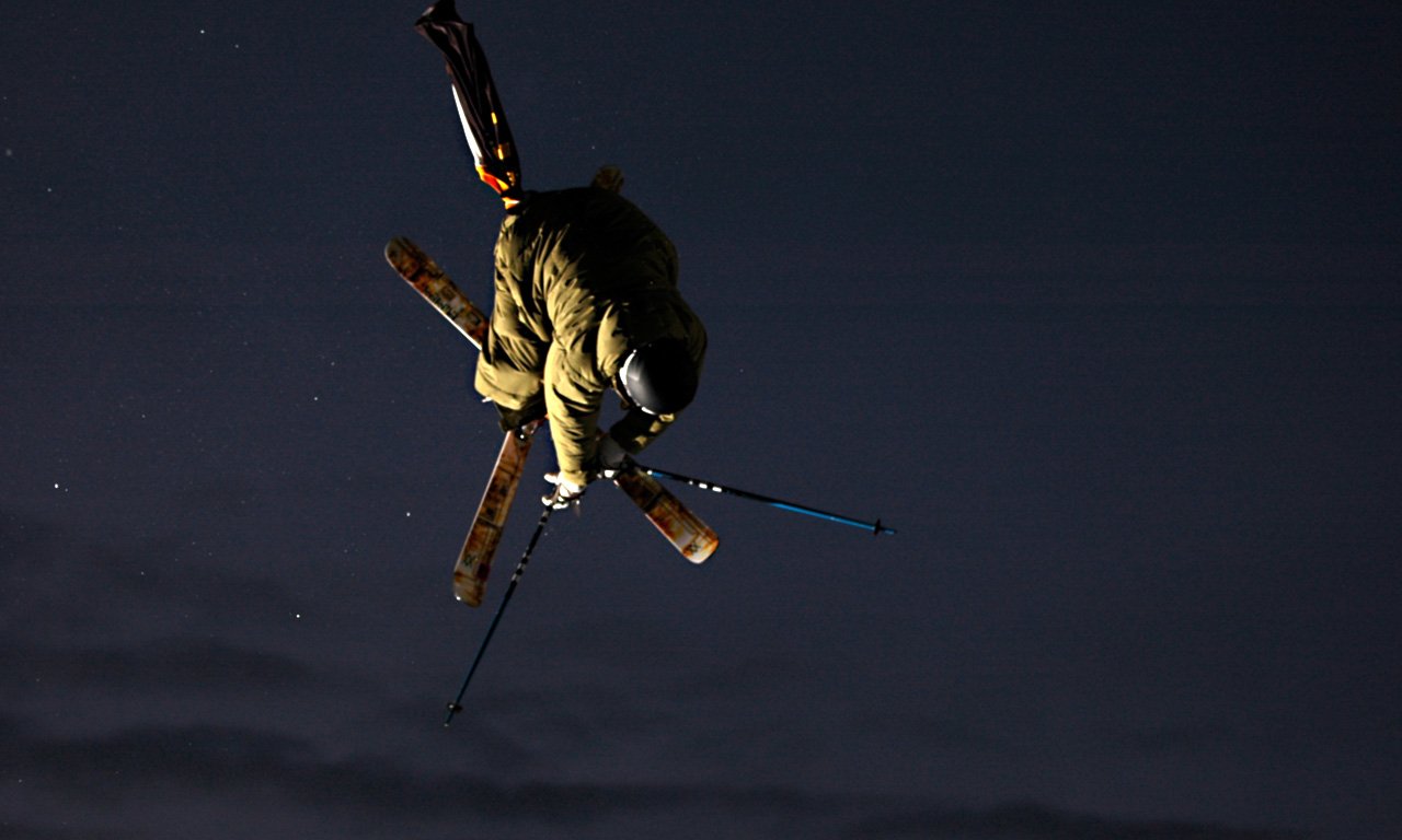 A skier performs a mid-air trick against a dark sky, showcasing skill and athleticism in this dynamic skiing sports scene.