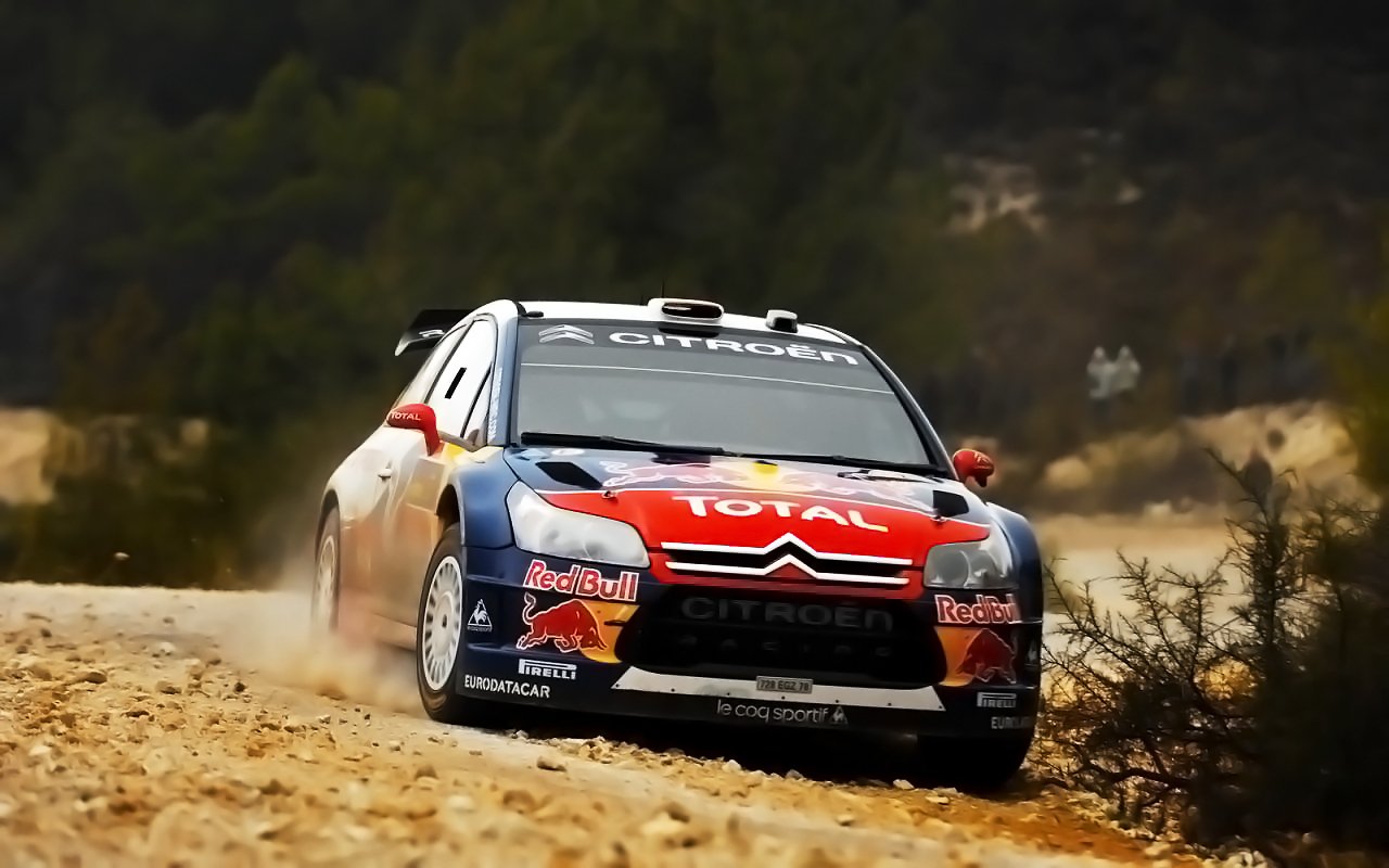 A rallye vehicle speeds along a dusty off-road trail, kicking up dirt as it navigates through a rugged terrain with sparse vegetation.