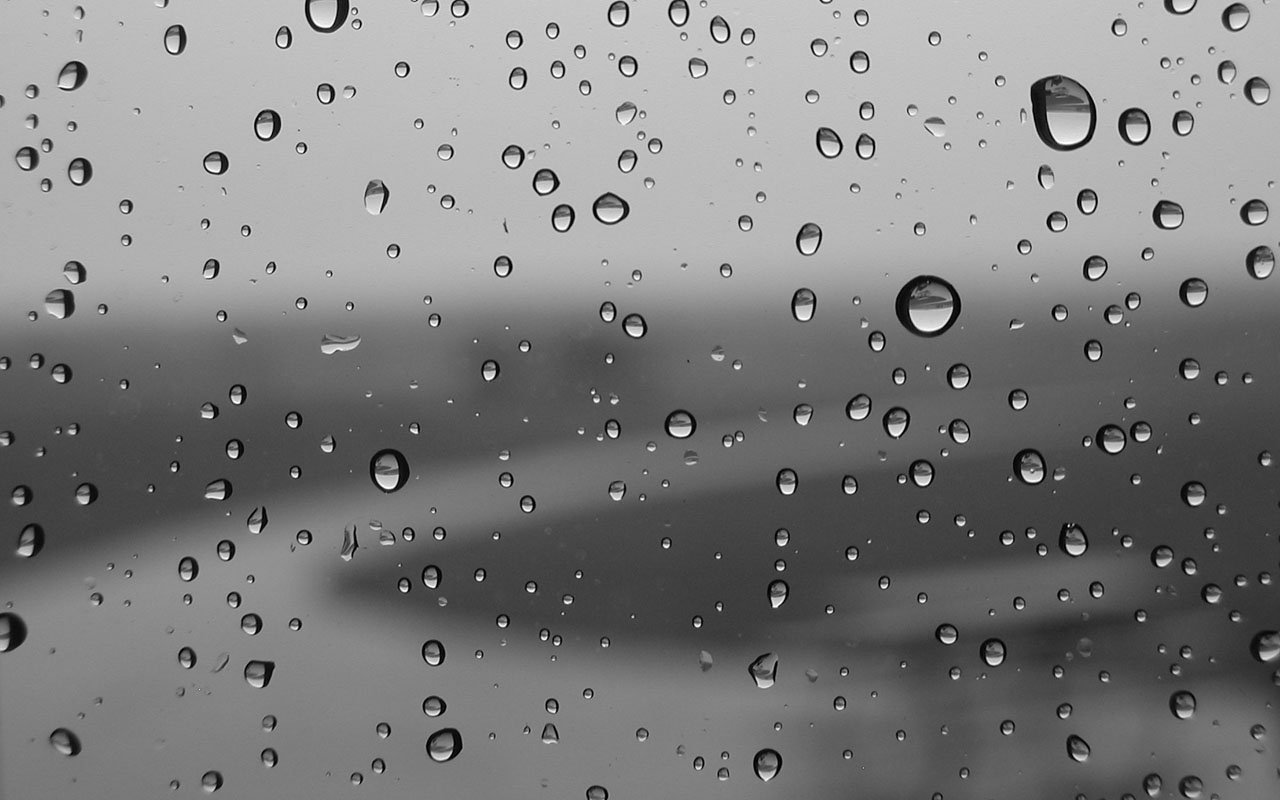 Droplets of water cling to a glass surface, creating a textured pattern against a blurred, monochromatic background. The photography captures a moment of serene beauty.