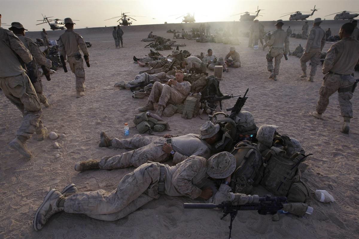 Soldiers resting on desert ground, equipped with military gear, while helicopters hover in the background, showcasing the intensity of a military operation.