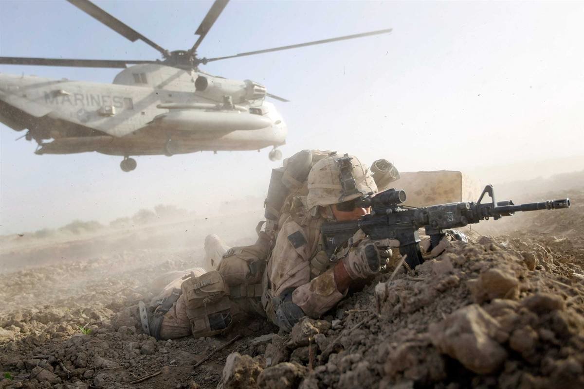 A U.S. Marine lies in a defensive position with a rifle, while a military helicopter hovers overhead in a dusty landscape, showcasing active ground operations.