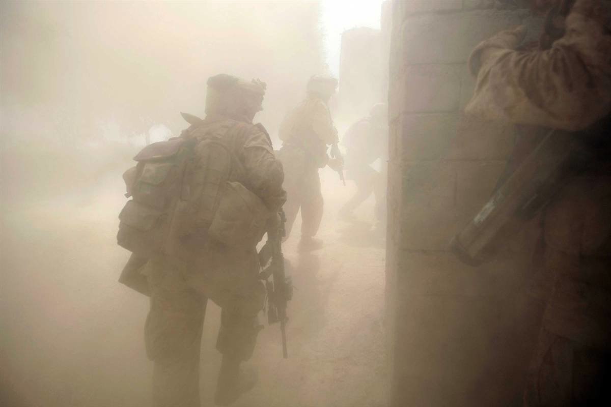 A group of soldiers navigates through a dusty environment, their figures partially obscured, as they approach a building during a military operation.