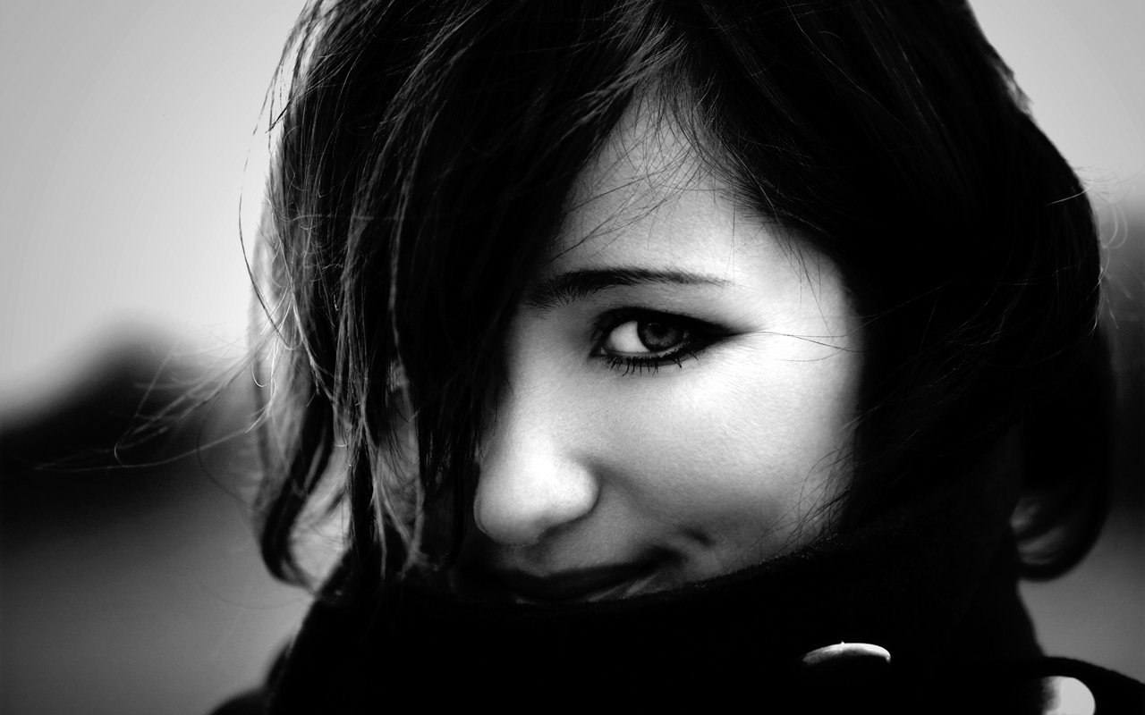 A pretty woman gazes playfully from behind her hair, with a slight smile, captured in a striking black and white portrait.