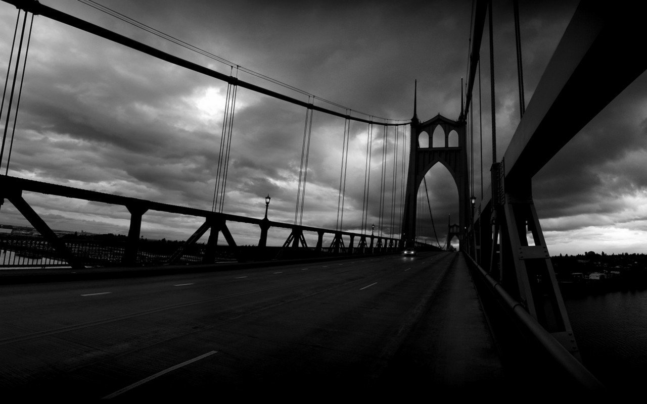 Black and white photo of the man-made St. Johns Bridge with dramatic clouds overhead, emphasizing its suspension structure and towering arch.