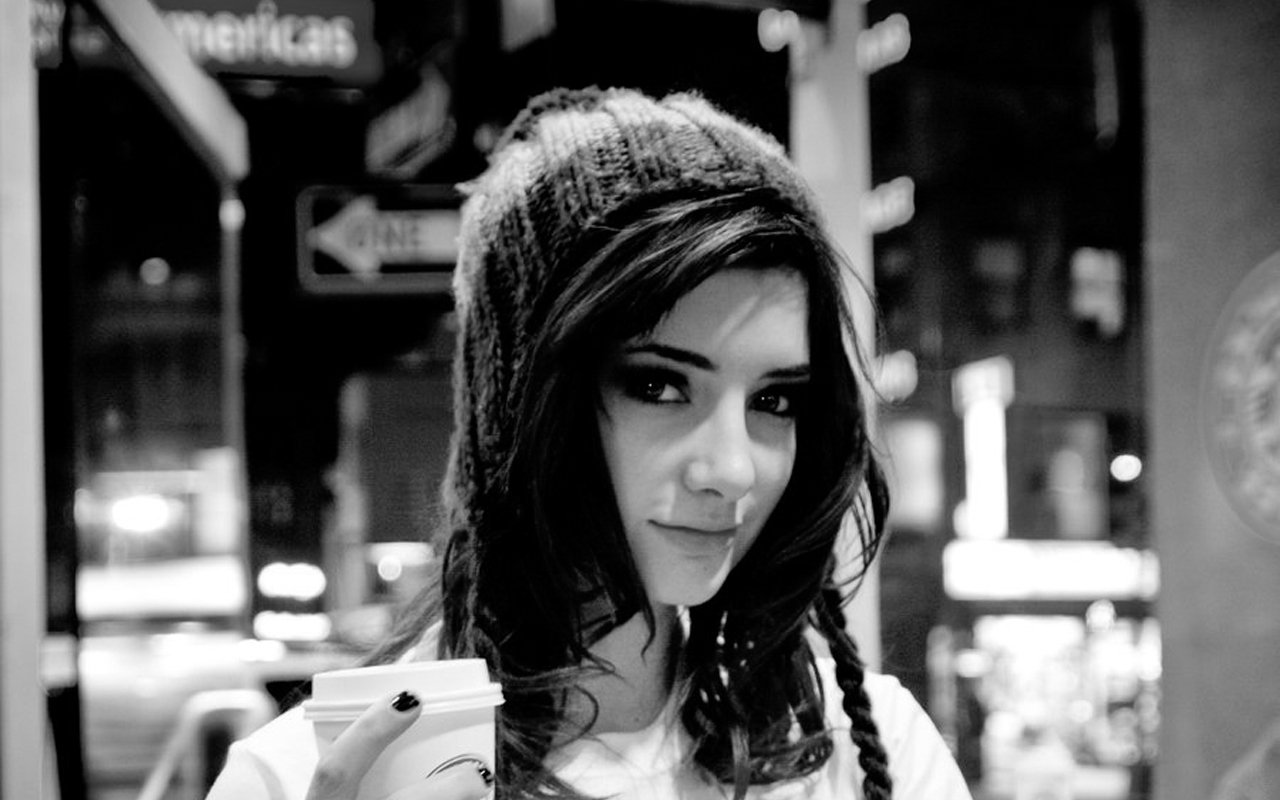A black and white portrait of Susan Coffey, a woman wearing a beanie, gazing confidently at the camera while holding a drink, showcasing an artistic vibe.