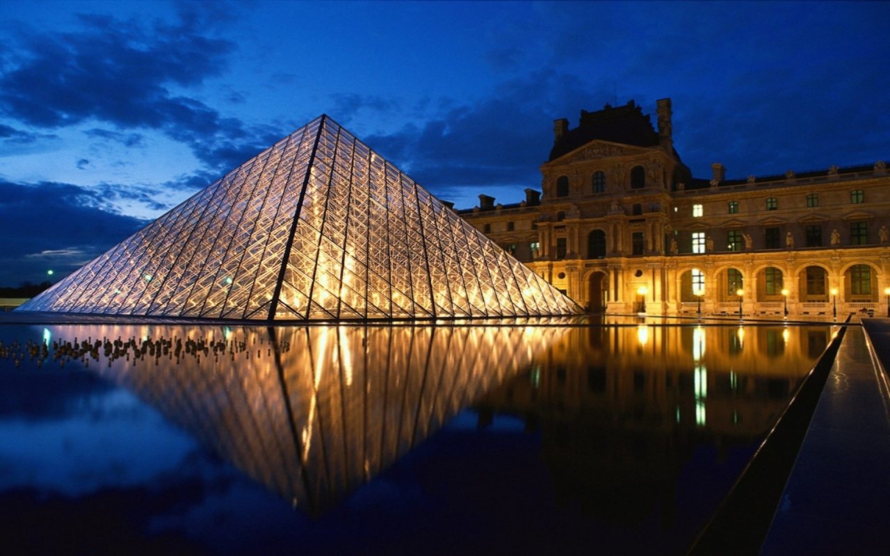 The Louvre Museum in Paris features its iconic glass pyramid, beautifully illuminated at dusk, reflecting in the tranquil waters nearby.