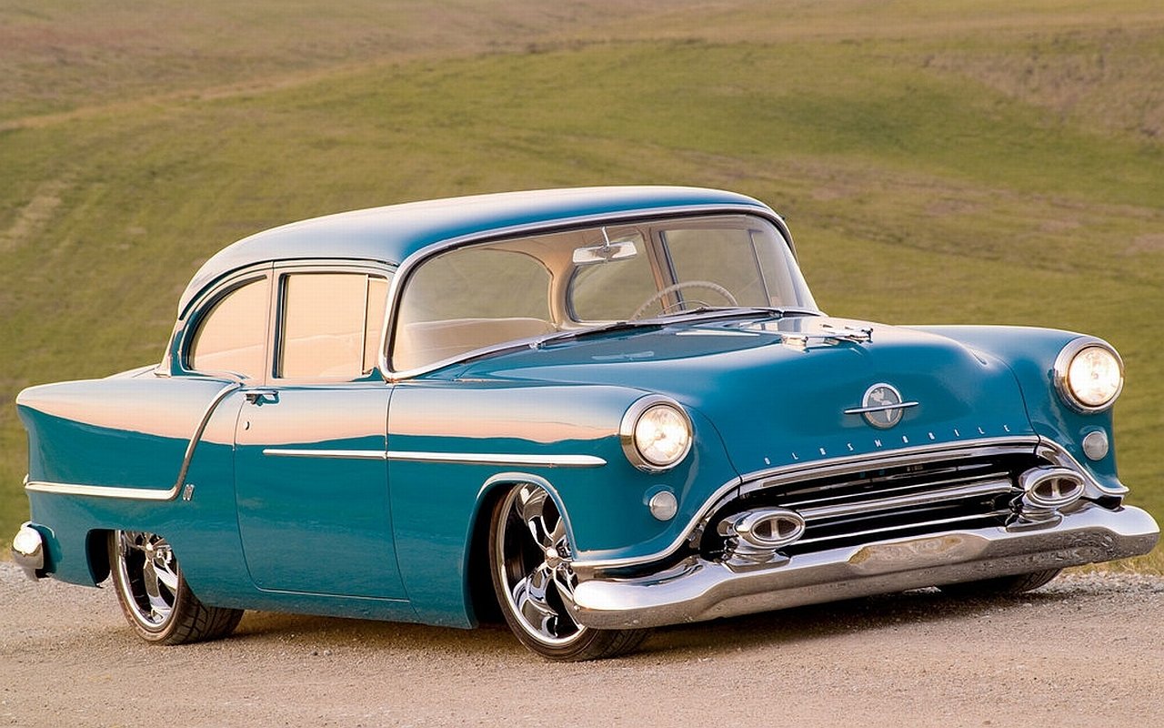 Teal vintage Oldsmobile coupe with chrome trim and custom wheels parked on a rural road.