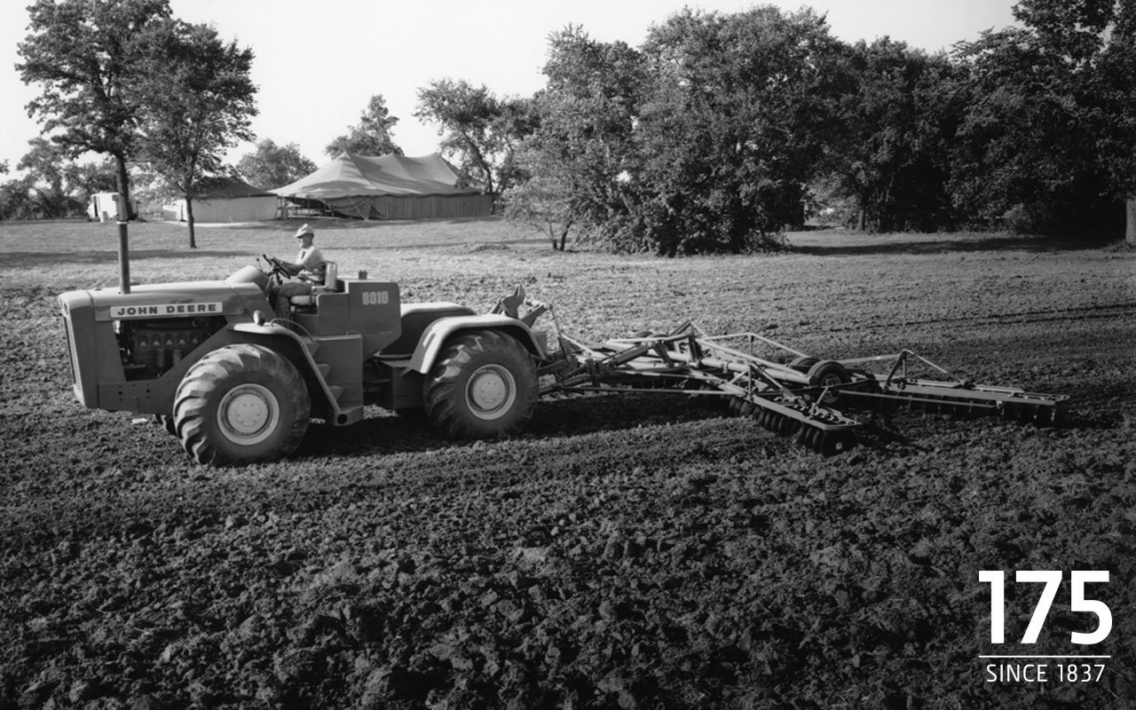 A John Deere vehicle with large tires is pulling farming equipment across a plowed field under a sky with scattered trees.