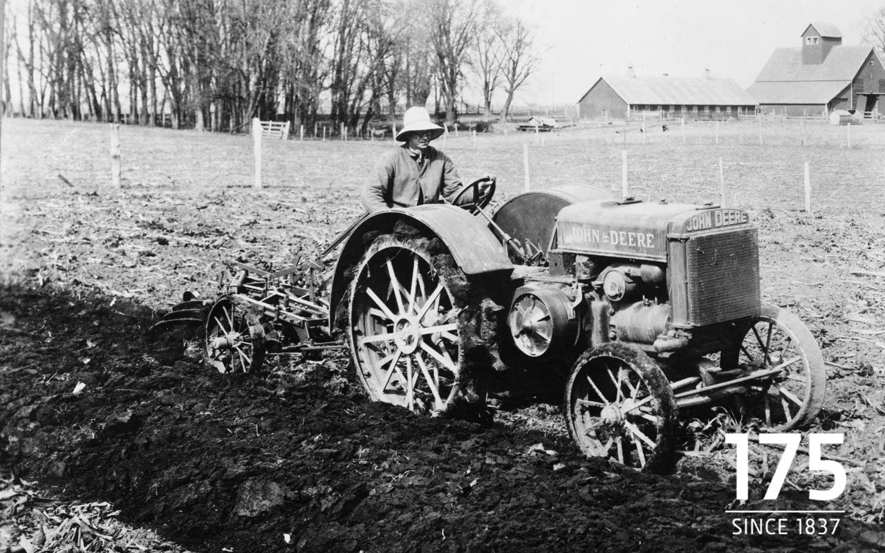 Vintage black and white photo of a John Deere vehicle plowing a field with a farmer seated on the tractor, rural buildings and leafless trees in the background.