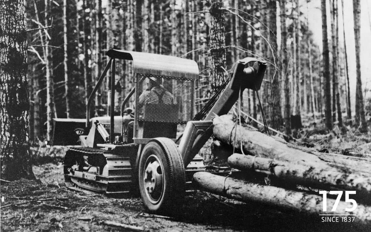 Black-and-white photo of a John Deere tracked vehicle transporting logs in a forested area.