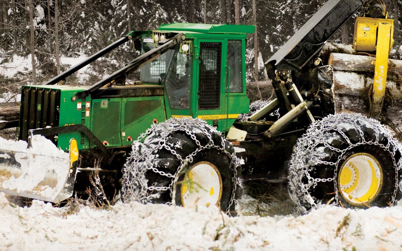 A John Deere vehicle equipped with large chain-covered tires moves through a snowy forest, designed for heavy-duty logging or forestry work.