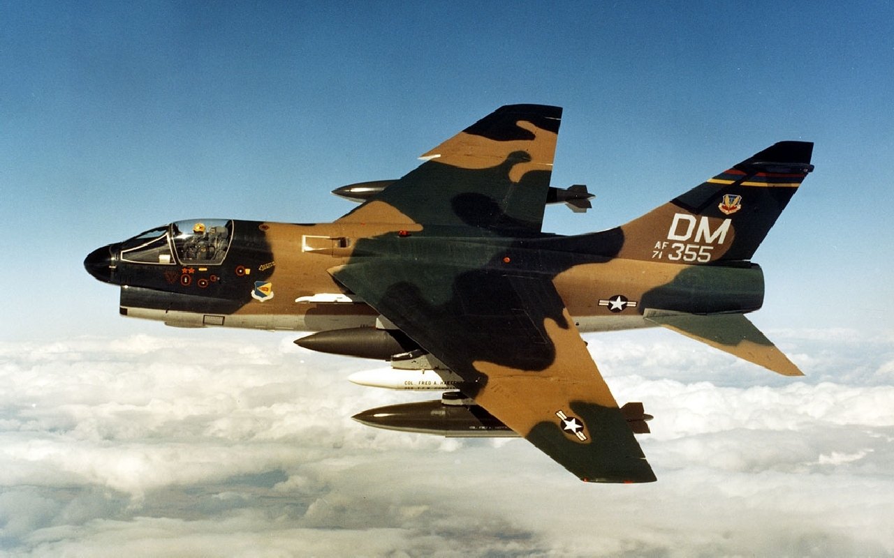 A military LTV A-7 Corsair II aircraft painted in camouflage flies above the clouds against a clear blue sky.