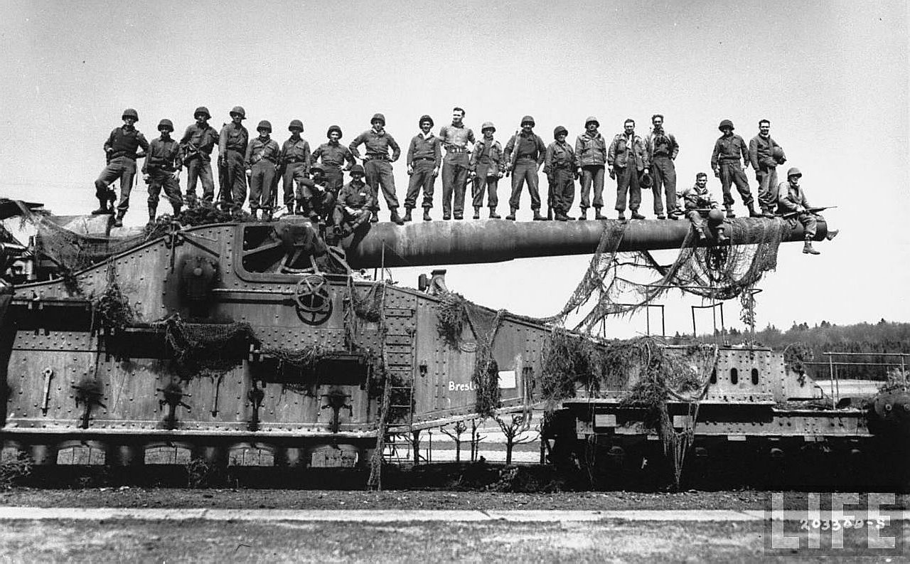 A group of soldiers poses atop a massive artillery piece, showcasing military equipment against a backdrop of expansive terrain. The image captures a moment of camaraderie and strength.