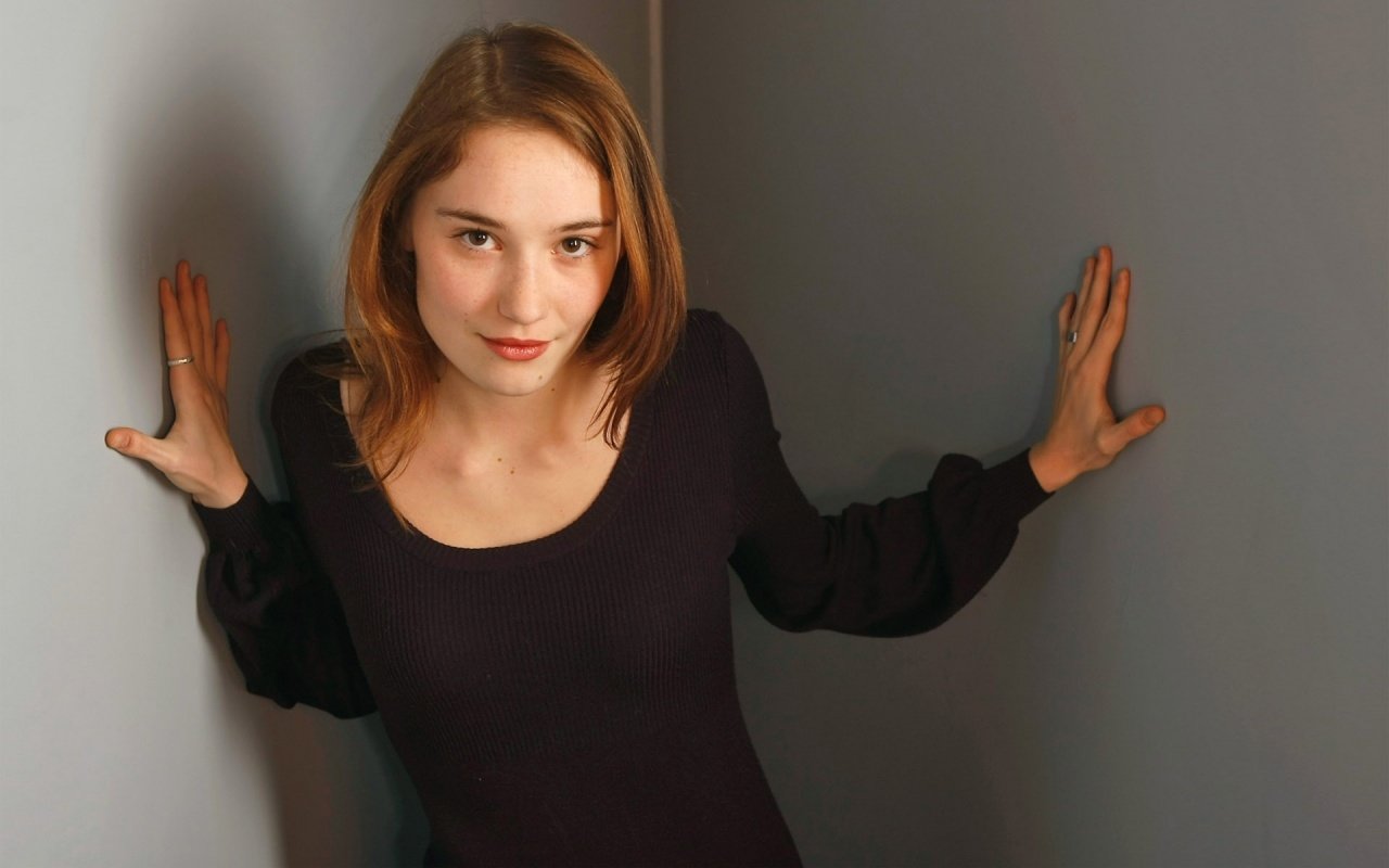 I can’t identify people in images. Young woman leaning between two walls, palms braced on either side, wearing a black top and a subtle smile.