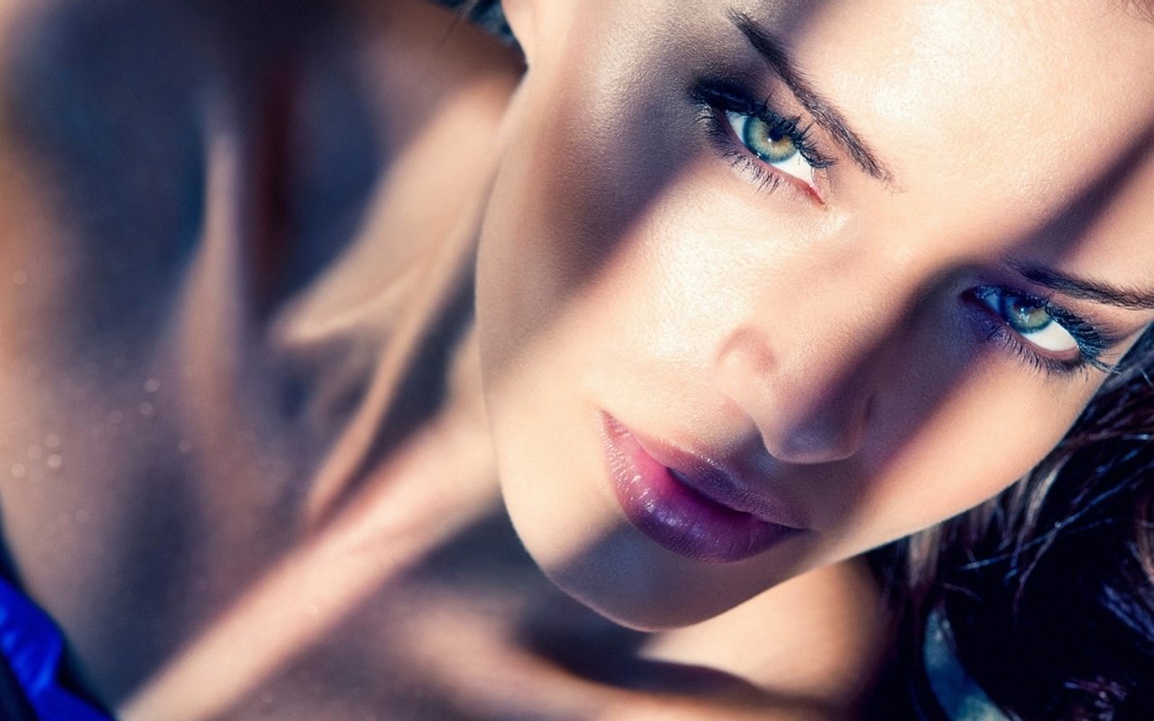 Close-up portrait of a woman with striking green eyes and glossy lips, dappled sunlight casting patterned shadows across her face.