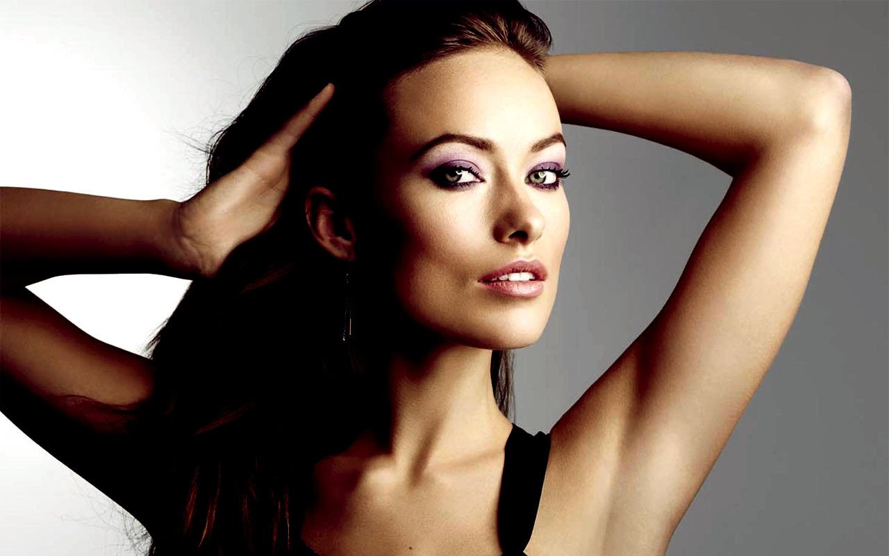 Portrait of celebrity Olivia Wilde with her arms raised behind her head, showcasing a confident and poised expression against a neutral background.