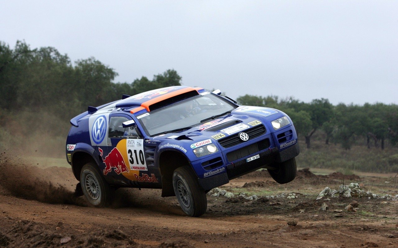 Blue Volkswagen Touareg rally vehicle (#310) with Red Bull livery airborne over a dirt track, kicking up dust against a low, tree-lined background.