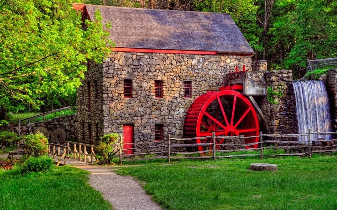 Stone man-made watermill with a large red waterwheel and flowing spillway, set among green lawn, trees, and a gravel path.