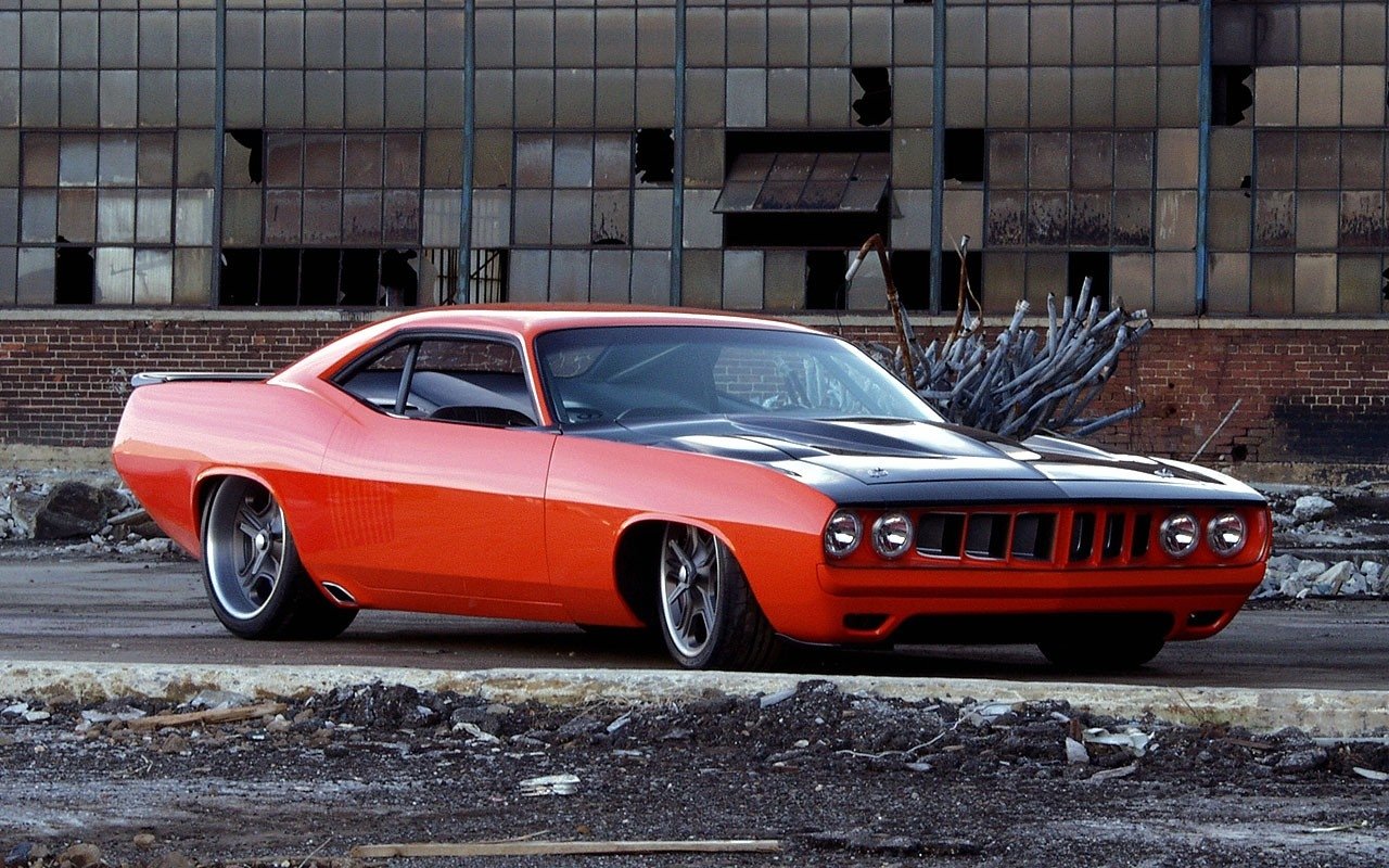 Orange Plymouth Barracuda vehicle with matte-black hood, low stance and chrome wheels parked in front of a weathered industrial building with broken windows.