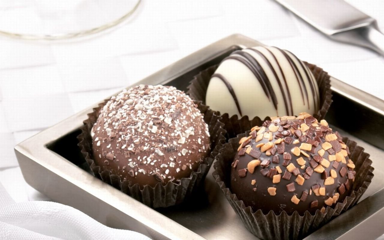 A silver tray holds three delectable chocolate treats: one coated in shredded coconut, another drizzled with dark chocolate stripes, and a third adorned with nuts.