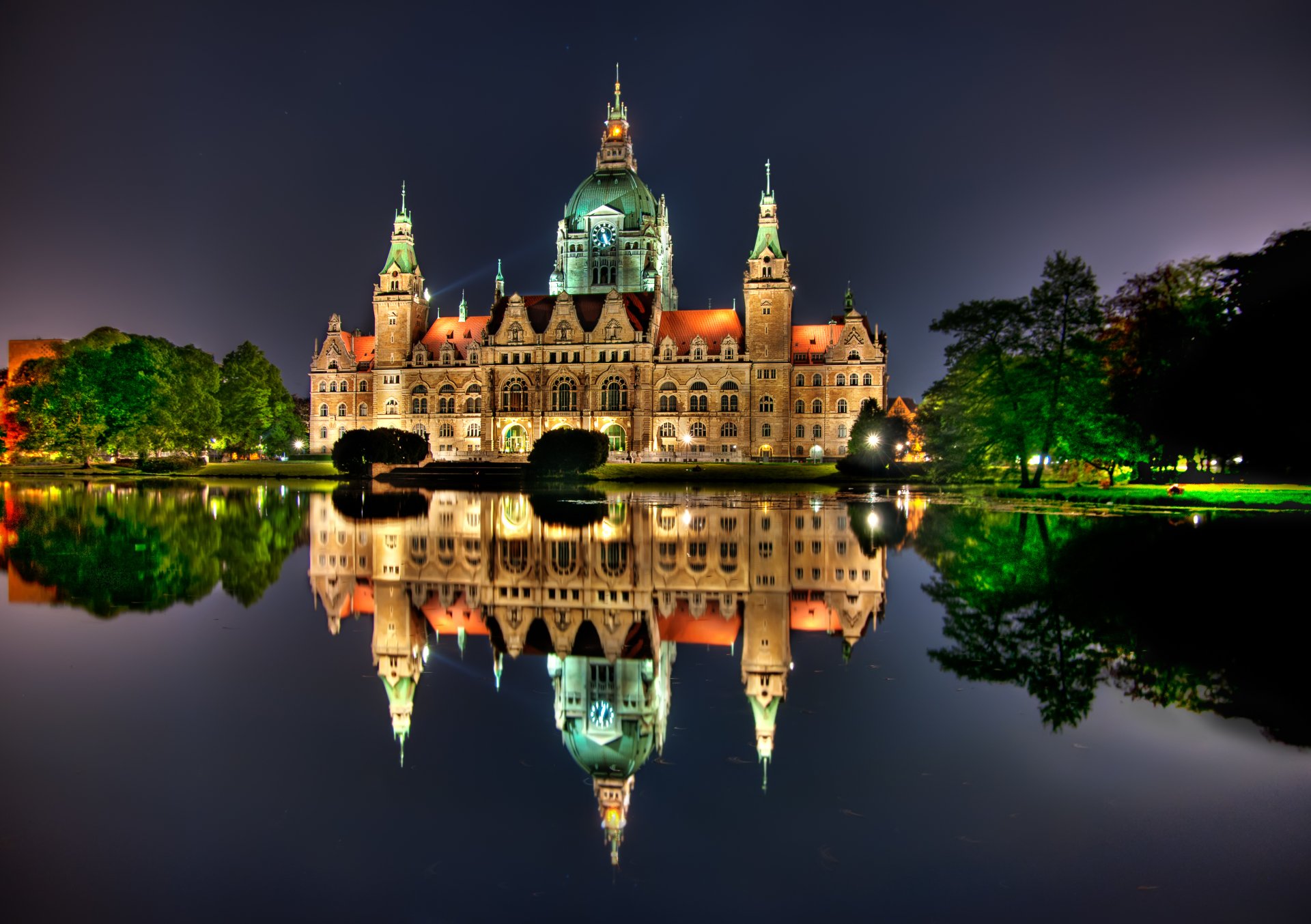 A stunning man-made building at night, reflecting beautifully in the water, surrounded by lush greenery and softly illuminated by lights.