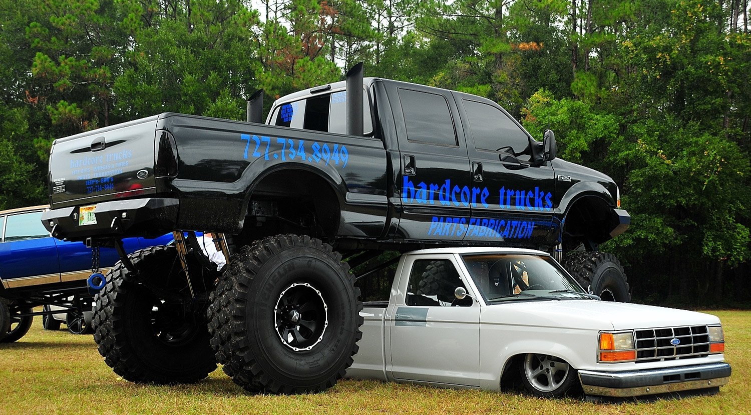 A large black truck with oversized tires towers over a smaller white truck parked underneath it, showcasing a striking contrast in vehicle size.