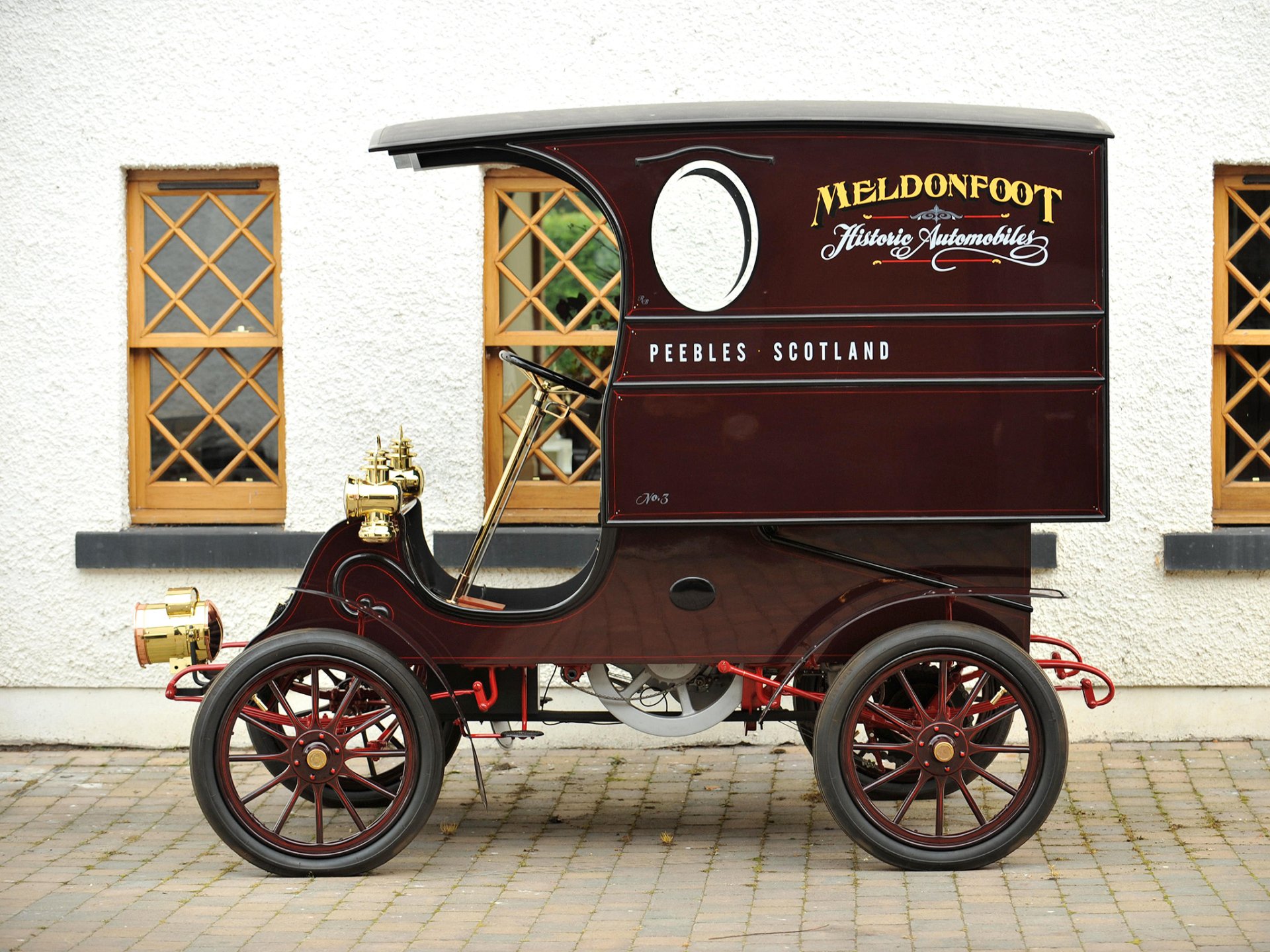 Maroon antique Cadillac delivery vehicle with covered cargo box and brass headlights parked in front of a white building with lattice windows.