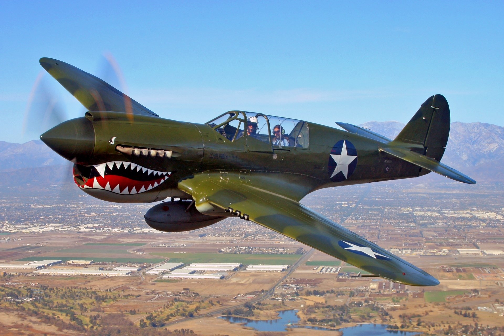 Military aircraft with a shark mouth design painted on the nose, flying over a landscape with mountains and fields under a clear blue sky.
