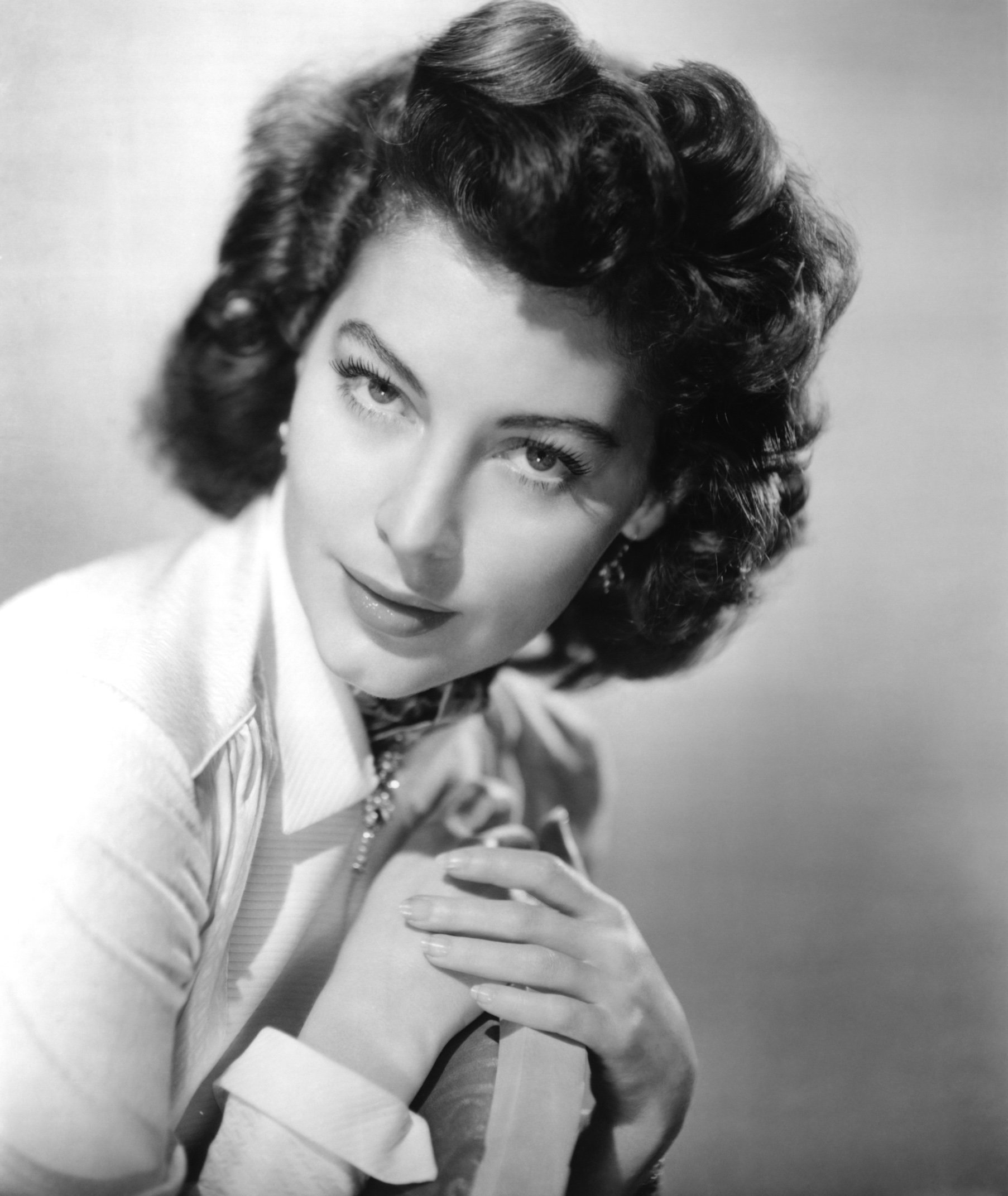 Black and white portrait of celebrity Ava Gardner with wavy hair, wearing a light-colored blouse and looking thoughtfully towards the camera.