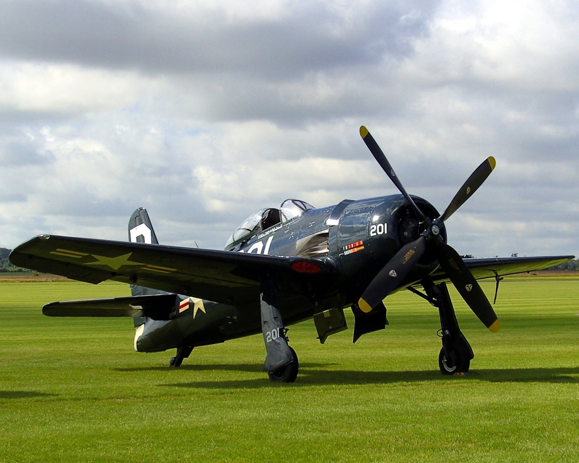 A vintage military aircraft with folded wings and a four-blade propeller parked on a grassy field under a cloudy sky.